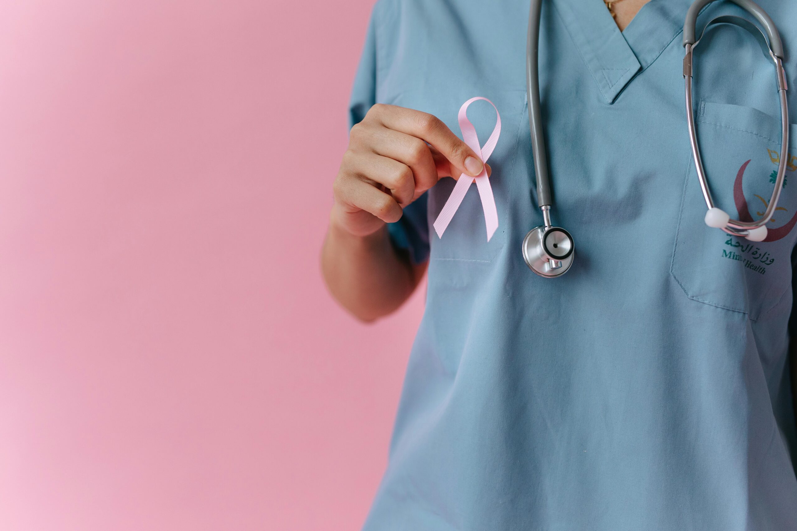 Healthcare worker in scrubs holds a pink breast cancer awareness ribbon symbolizing support.